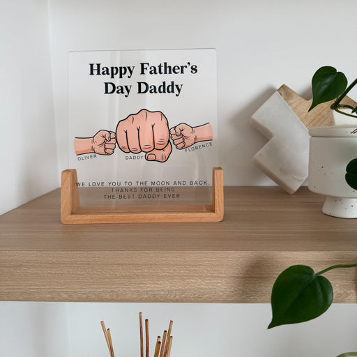 A Father's Day glass sign displayed on a wooden shelf. The sign features three illustrated hands clasped together with the text 'Happy Father's Day Daddy' and personalized names 'Oliver', 'Daddy', and 'Percy' above it.