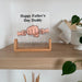 A Father's Day glass sign displayed on a wooden shelf. The sign features three illustrated hands clasped together with the text 'Happy Father's Day Daddy' and personalized names 'Oliver', 'Daddy', and 'Percy' above it.