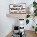 A wooden sign reading 'GRANDAD'S Whiskey Bar' with 'COLD DRINKS & GOOD TIMES' displayed on a light wood floating shelf above a bar counter. The sign features a black border and black lettering on a light natural wood background.