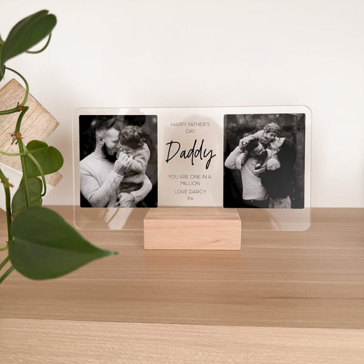 A personalized acrylic photo plaque on a small wooden stand, displayed on a light wood shelf. The plaque features three sections: two black-and-white photos of a man holding a child, and a central white section with the text 'Happy Father's Day Daddy YOU ARE ONE OF MY BEST LOVE GADGETS'.