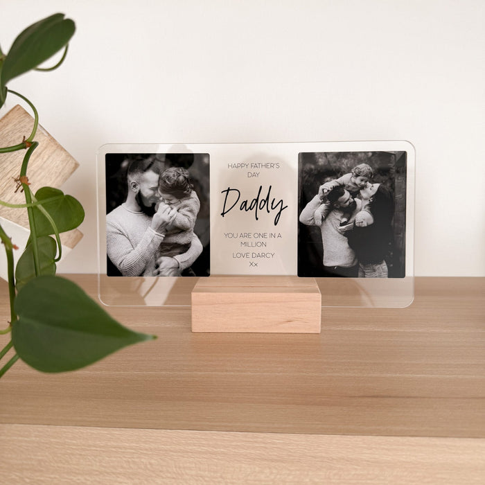 A personalized acrylic photo plaque on a small wooden stand, displayed on a light wood shelf. The plaque features three sections: two black-and-white photos of a man holding a child, and a central white section with the text 'Happy Father's Day Daddy YOU ARE ONE OF MY BEST LOVE GADGETS'.