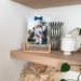 A framed custom photo print with a wooden stand on a light wood floating shelf, styled with a black and white striped vase, a white ceramic jar, and dried flowers below.