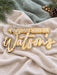 Layered family name sign in white, gold mirror and frosted acrylic, lying flat on linen to show the 2 hanging holes