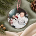 Baby photo ornament in glossy white ceramic showing a baby in white beside a toy reindeer, on red and white twine